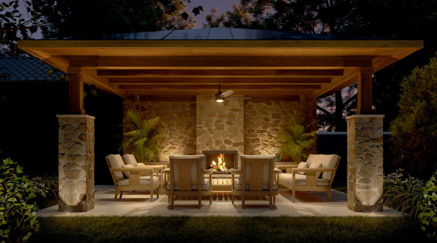 Outdoor living area centered around a fireplace under a wood-beam-covered patio sustained by flagstone and concrete columns.