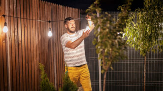 Man hanging outdoor string lights next to a tree and backyard fence.