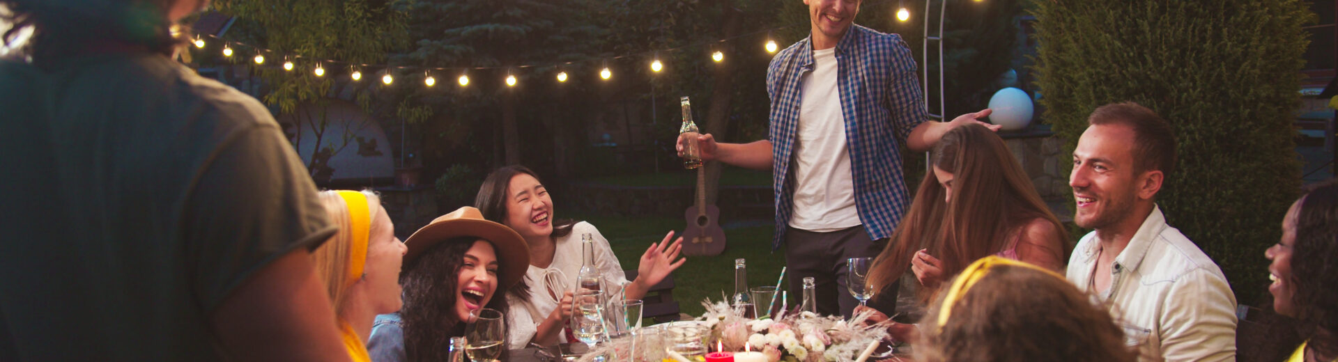 Exterior party lights hanging over a social gathering surrounding a table full of food.