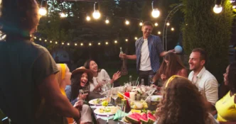 Exterior party lights hanging over a social gathering surrounding a table full of food.