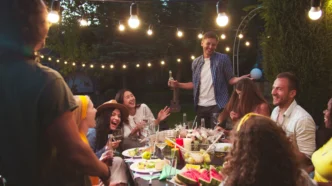 Exterior party lights hanging over a social gathering surrounding a table full of food.