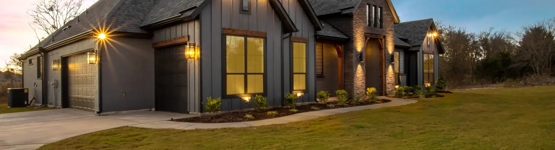 Angled view of upper middle class farm house with outdoor lighting at dusk.