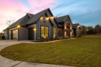 Angled view of upper middle class farm house with outdoor lighting at dusk.