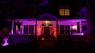 Front of two-story home illuminated with purple landscape lighting and decorative gravestones and ghosts festooned throughout for Halloween vibe.