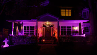 Front of two-story home illuminated with purple landscape lighting and decorative gravestones and ghosts festooned throughout for Halloween vibe.