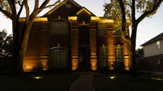 Uplights and inground well lights illuminating an upper middle class brick house at dusk.