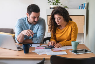 Young couple analyzing their bills to budget expenses strewn out in front of them at a table with a laptop.