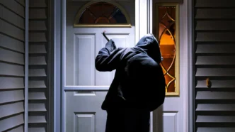 Burglar dressed in all black attempting to break into a home but deterred by a bright motion sensor light near entryway.