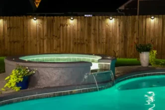 A backyard swimming pool and jacuzzi hot tob at night with solar lights around the fence for privacy and illumination.