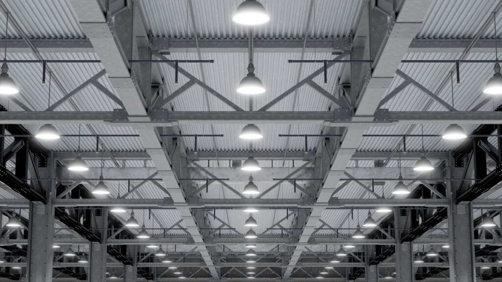 Rows of bright pendant lights hanging between steel rafters attached to the ceiling of a warehouse or factory.