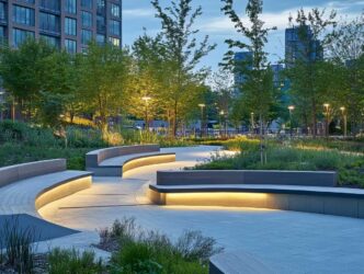 Urban park with strip lights tucked under benches and lights illuminating green trees with a cityscape in the background.
