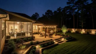 Residential backyard at night complete with a deck-style patio illuminated with patio string lights and path lights for guiding traffic as well as uplights illuminating a wood fence perimeter.