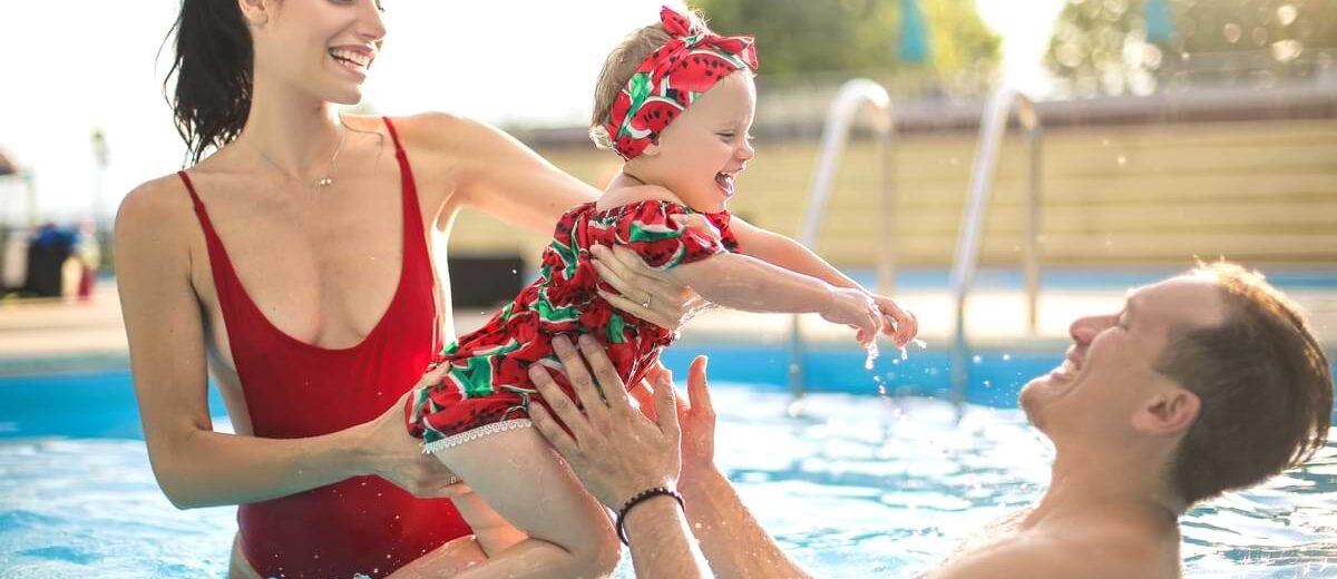 Toddler girl in a red bathing suit jumping into her dad's arms with her mom in a swimming pool.