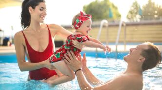 Toddler girl in a red bathing suit jumping into her dad's arms with her mom in a swimming pool.