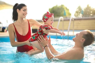 Toddler girl in a red bathing suit jumping into her dad's arms with her mom in a swimming pool.