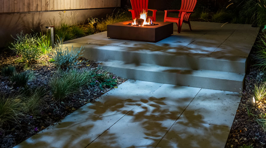 Tree leaf shadows on the pavement of a sidewalk leading to a patio seating area with a firepit and two red Adirondack chairs.
