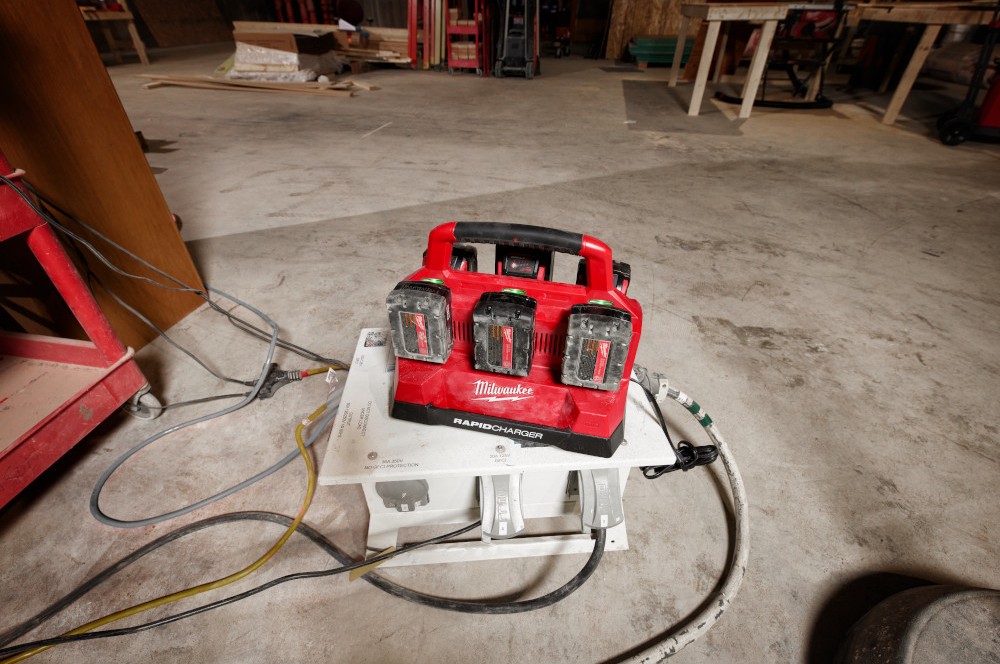 Milwaukee Six-Bay Rapid Charger charging six Milwaukee batteries on the concrete ground of a jobsite.
