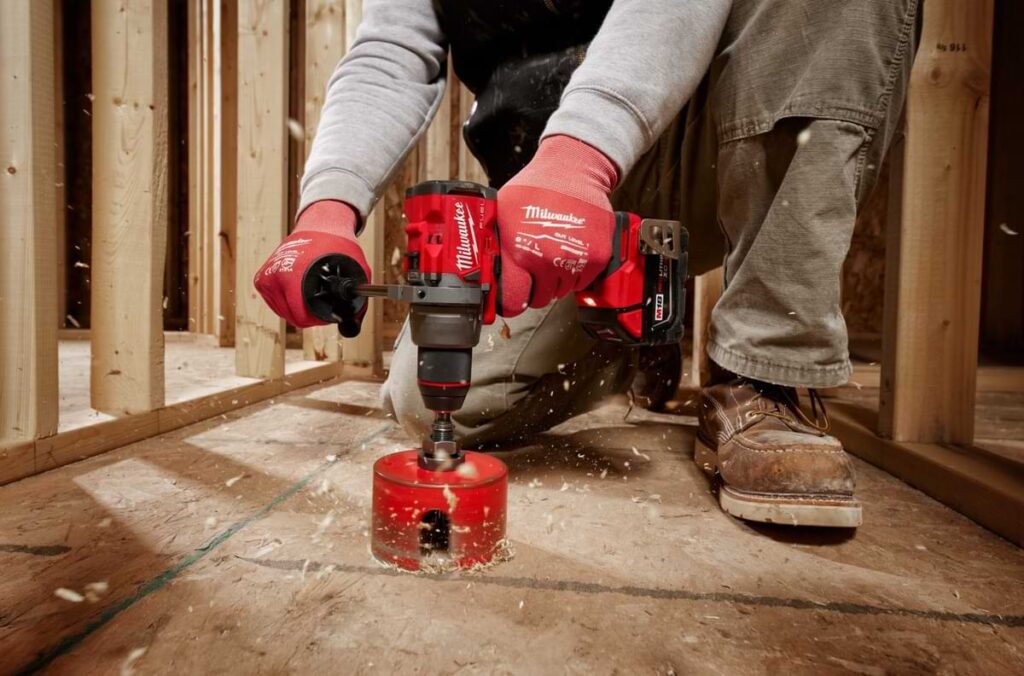 Person using a Milwaukee Tool power drill to drill through plywood with wood framing in the background.