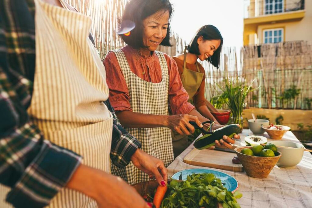 Three people, including a mother and adult daughter, preparing vegetables in an outdoor kitchen under the shining sun. 