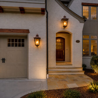 Front of white brick home with two outdoor coach lights, one above the doorway arch and one to the right of the garage door.